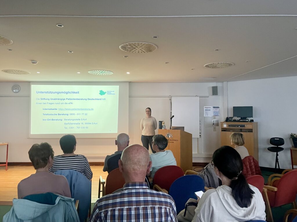 Martina steht vor einer Gruppe älterer Menschen, die auf Stühlen sitzen. Hinter ihr ist eine Präsentation an die Wand projiziert.