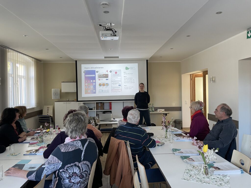 Martina steht vor einer Gruppe älterer Menschen. Hinter ihr ist eine Leinwand mit einer Präsentation.