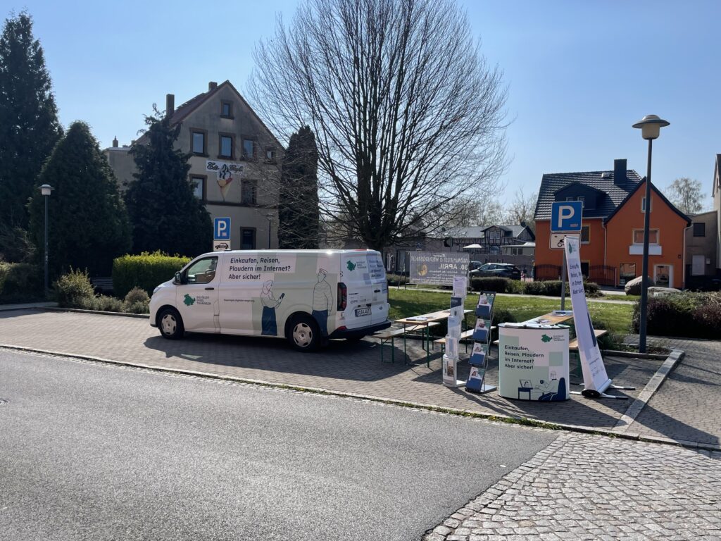 Das Infomobil und der Infostand des Projekts stehen auf einem großen Platz. Im Hintergrund ist blauer Himmel und ein Baum zu sehen.