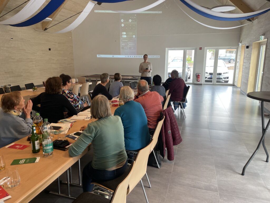 Ein Raum mit einem langen Tisch, an dem ältere Menschen sitzen. Martina steht vor dem Tisch und spricht zu den Anwesenden.