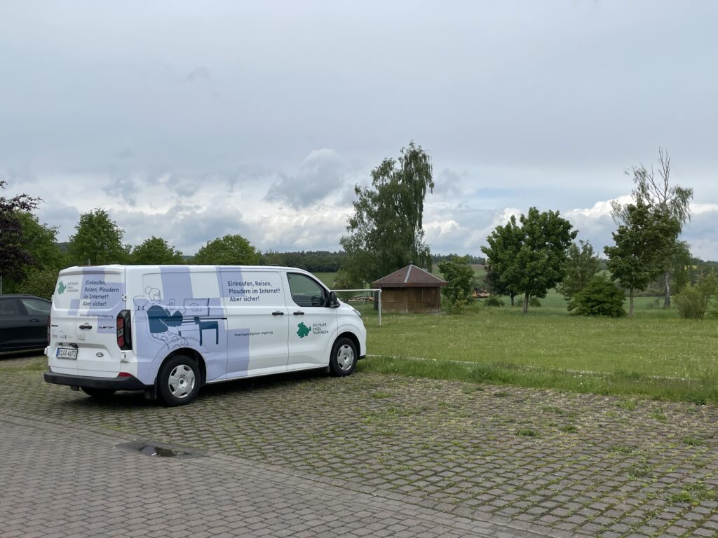 Das Infmobil steht auf einem leeren Parkplatz. Im Hintergrund ist Natur zu sehen.