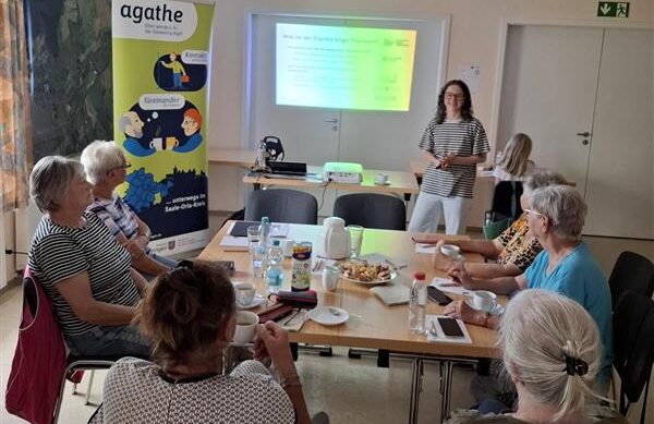 Sophie steht vor einer Gruppe älterer Menschen. Hinter ihr ist eine Präsentation an die Wand projiziert.