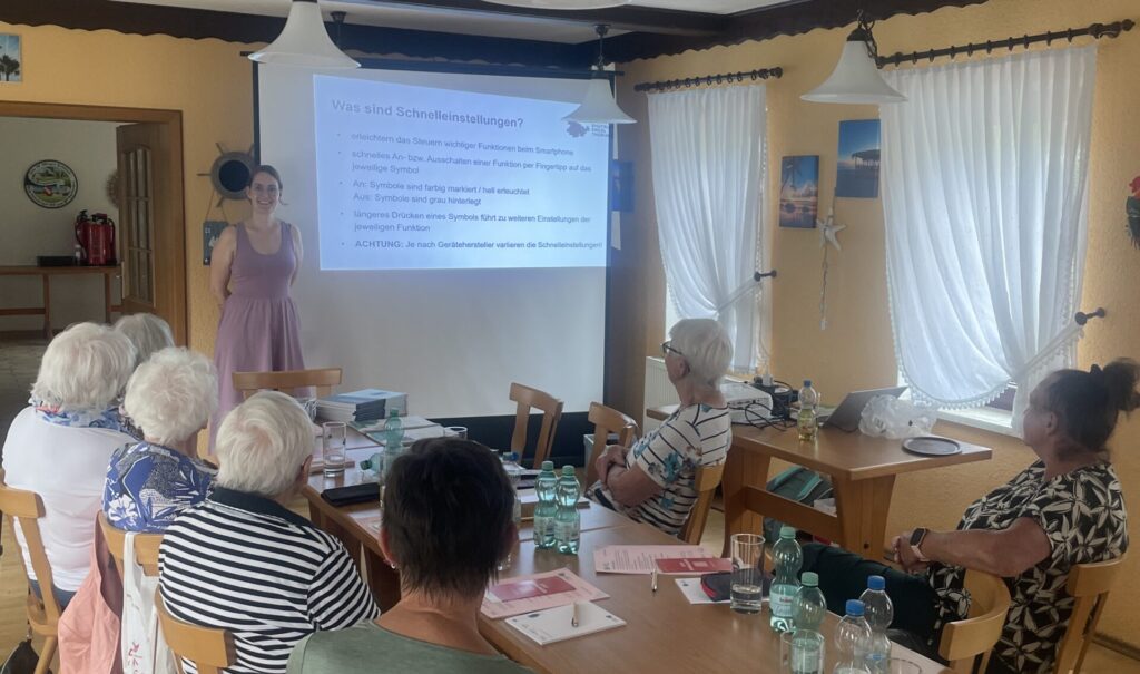 Martina steht vor einer Gruppe älterer Menschen, die an einem Tisch sitzt. Hinter ihr ist eine Präsentation an die Wand projiziert.