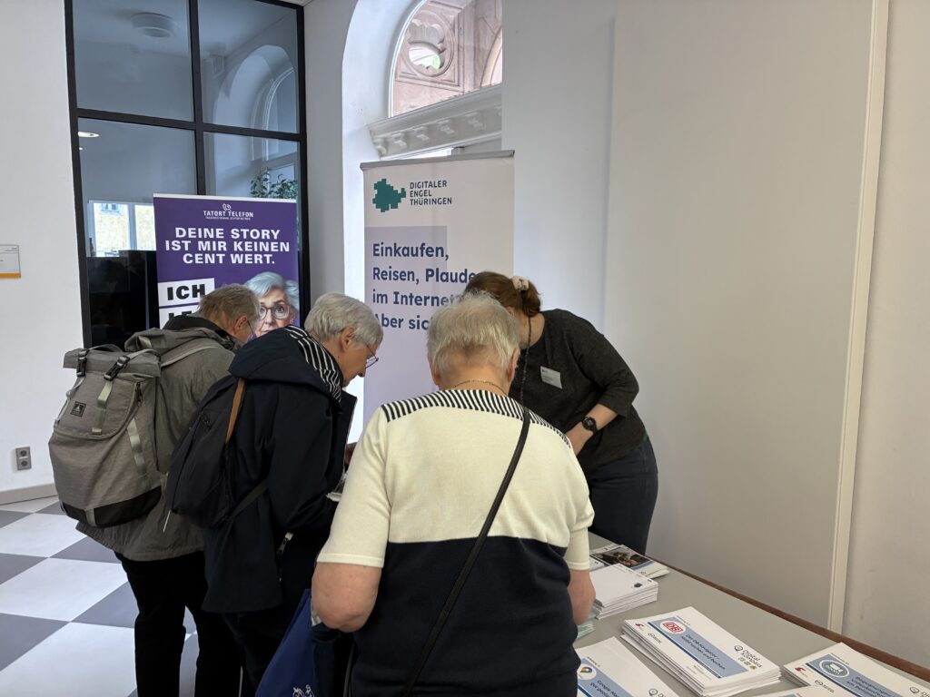 Drei ältere Personen stehen an einem Infostand mit Flyern, im Hintergrund zwei aufrechte Werbebanner in einem hellen Raum mit Fenster und Bogen.