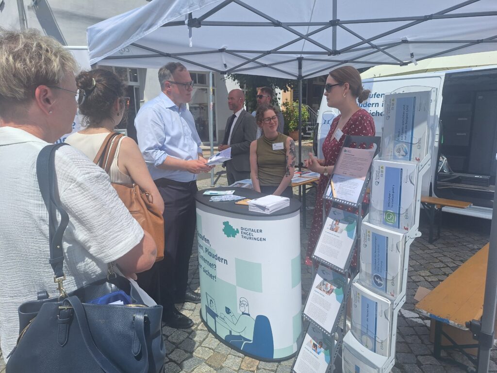 Menschen stehen an einem Informationsstand unter einem weißen Pavillon mit Flyern und Broschüren auf einem Pflasterplatz