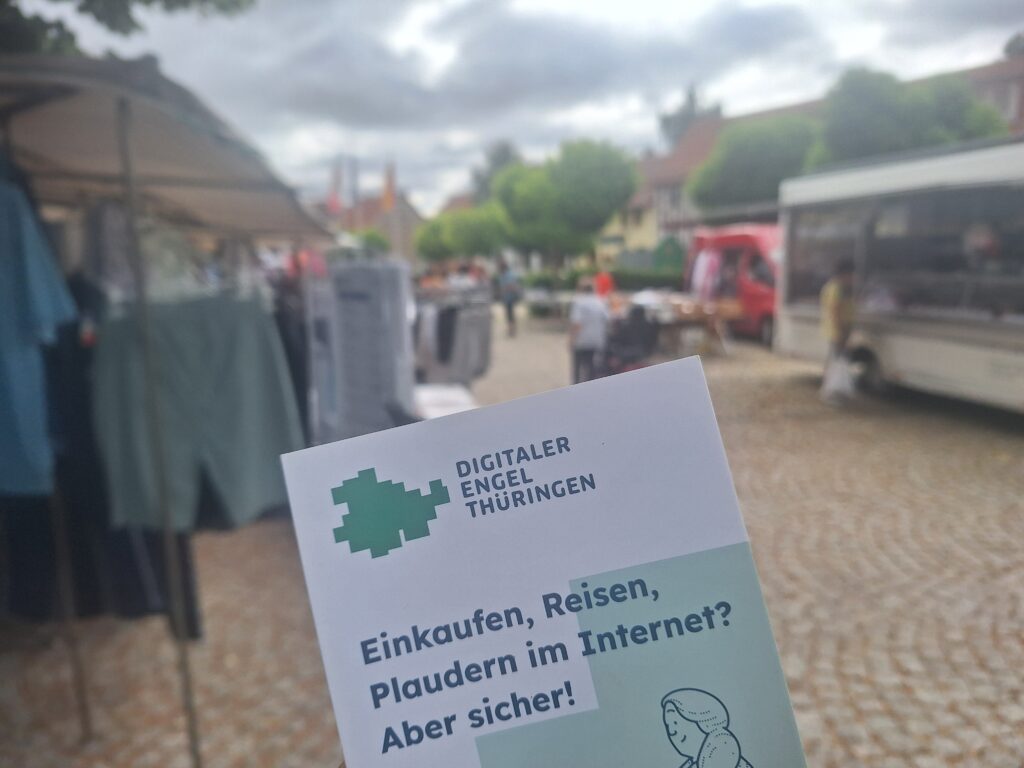 Im Vordergrund: Ein Flyer des Digitalen Engels Thüringen. Dahinter ist verschwommen ein Marktplatz mit Ständen und Häusern zu sehen.
