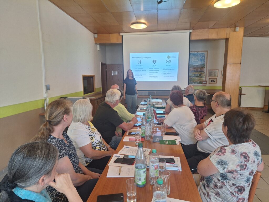 Martina steht vor einer Gruppe älterer Menschen, die an einem langen Tisch sitzt. Hinter Martina ist eine Leinwand mit einer Präsentation zu sehen.