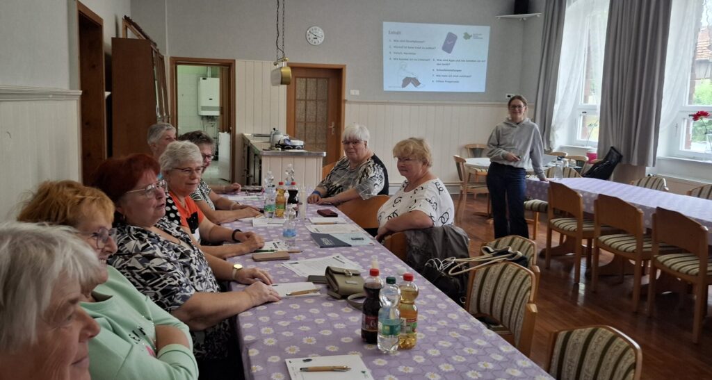 Sophie steht vor einer Gruppe älterer Menschen, die an einem Tisch sitzt. Hinter ihr ist eine Präsentation an die Wand projizuert.