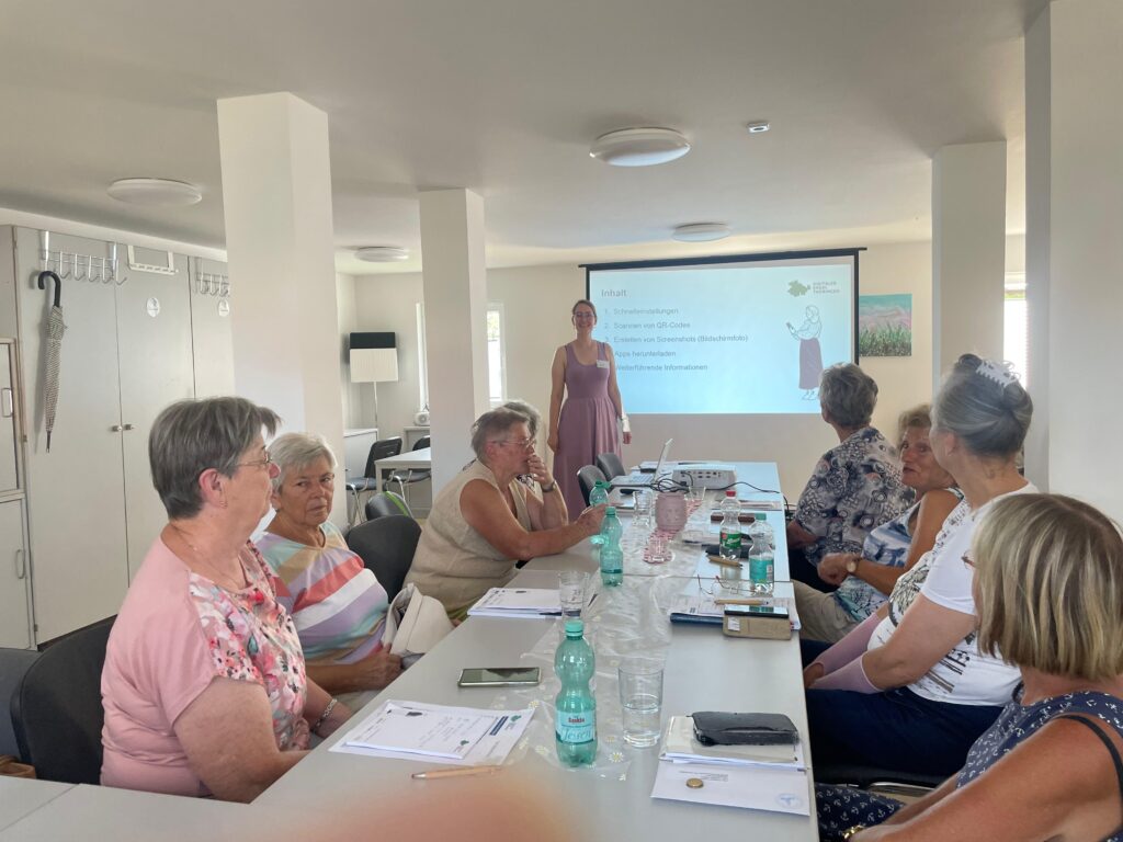 Martina steht in einem Raum mit einer Gruppe älterer Menschen, die an einem Tisch sitzt. Hinter MArtina ist eine Präsentation an die Wand projiziert.
