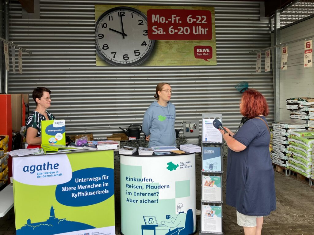 Martina steht am Stand des Digitalen Engels Thüringen. Im Hintergrund ist ein Supermarkt zu sehen. Eine Person steht vor dem Stand und hält Martina ein Mikrofon hin. Neben Martina steht eine weitere PErson.