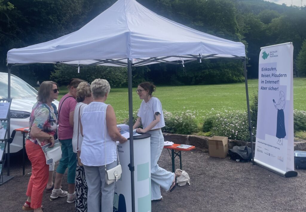 Sophie steht am Stand des Digitalen Engels Thüringen und spricht mit älteren Personen, die am Stand stehen.