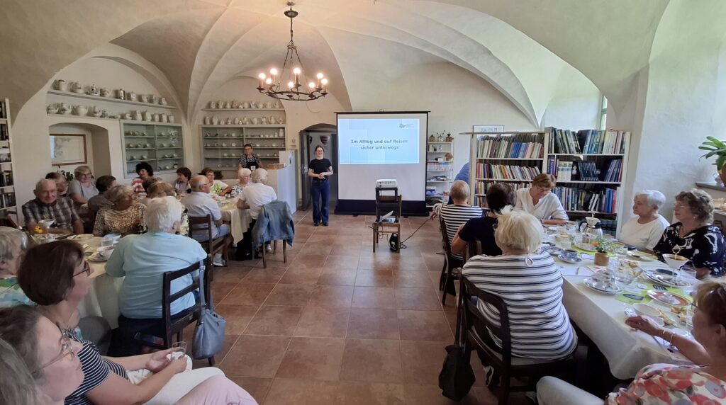 Sophie steht in einem großen gewölbten Saal vor einer Gruppe älterer MEnschen. Hinter ihr ist eine Präsentation an die Wand geworfen.