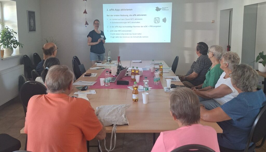 Martina steht in einem Raum mit einer Gruppe älterer Menschen, die an einem Tisch sitzt. Hinter MArtina ist eine Präsentation an die Wand projiziert.