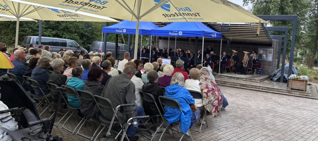 Ältere Menschen sitzen auf Stühlen unter Schirmen. Vor ihnen sitzt ein Orchester, das auf Instrumenten spielt.