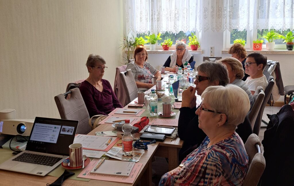 Ein langer Tisch, an dem mehrere ältere Menschen sitzen und nach vorne schauen. Auf dem Tisch sind Unterlagen und Geräte verteilt.