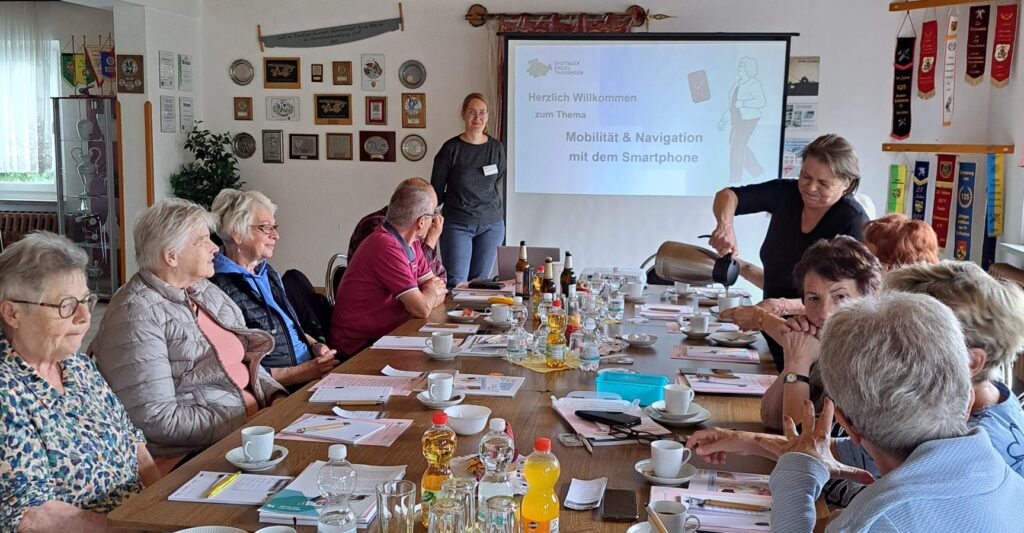 Mehrere ältere Menschen sitzen an einem langen Tisch mit Kaffeetassen und Unterlagen, eine Frau gießt Kaffee, im Hintergrund steht eine weitere Frau vor einer Leinwand mit der Projektion 'Herzlich Willkommen zum Thema Mobilität & Navigation mit dem Smartphone'