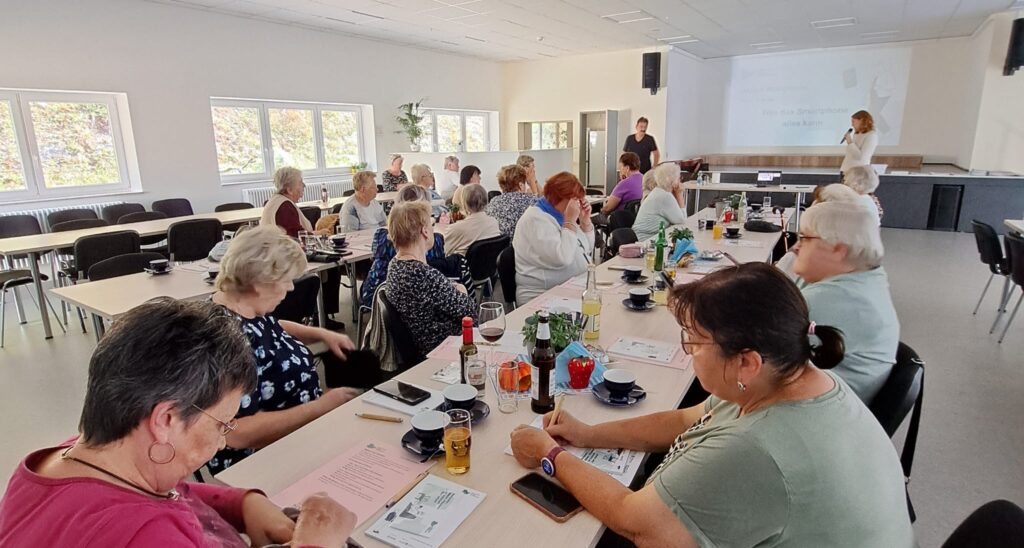 Viele ältere Menschen sitzen an einem langen Tisch und beugen sich über ihre Smartphones. Martina steht am Ende des Tischs.