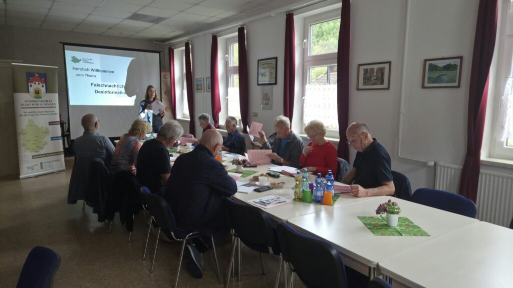 Martina steht am Ende eines langen Tischs. Am Tisch sitzen ältere Menschen. Hinter Martina ist eine Leinwand mit einer Präsentation zu sehen.