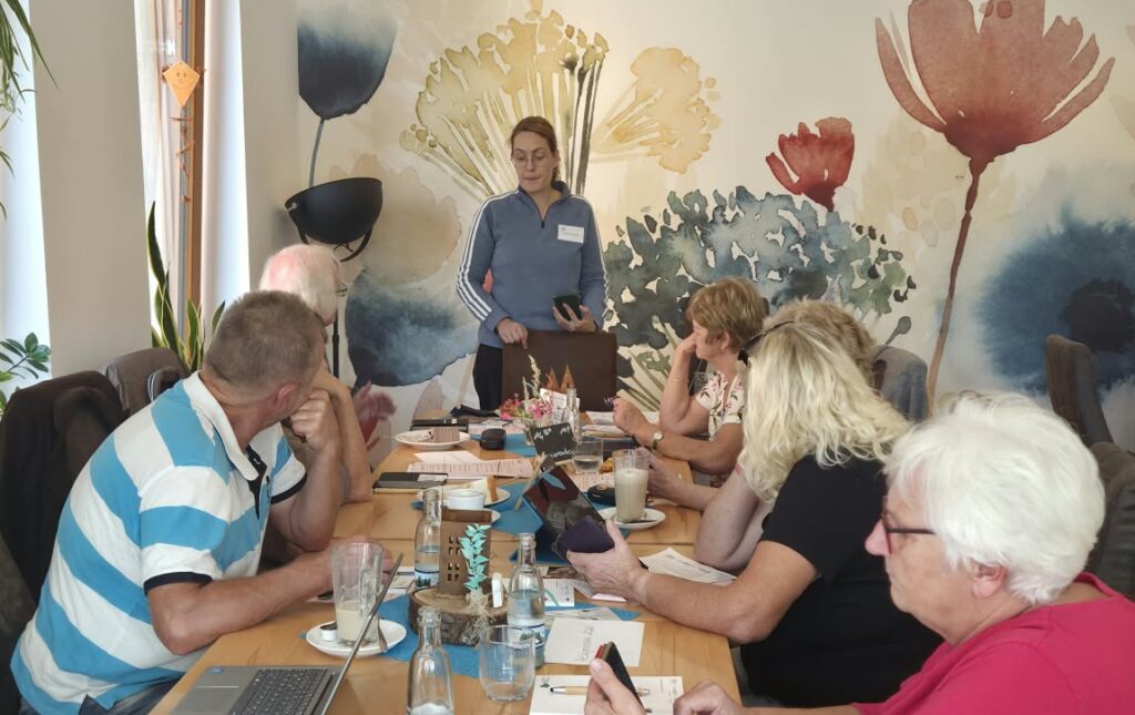 Martina steht vor einer Gruppe älterer Menschen, die an einem Tisch sitzt. Sie halten Smartphones in der Hand.