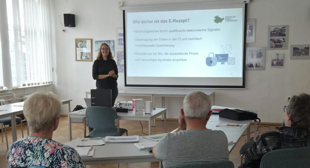 Martina steht vor einer Leinwand in einem großen Raum. Vor ihr sitzen ältere Menschen an einem Tisch.