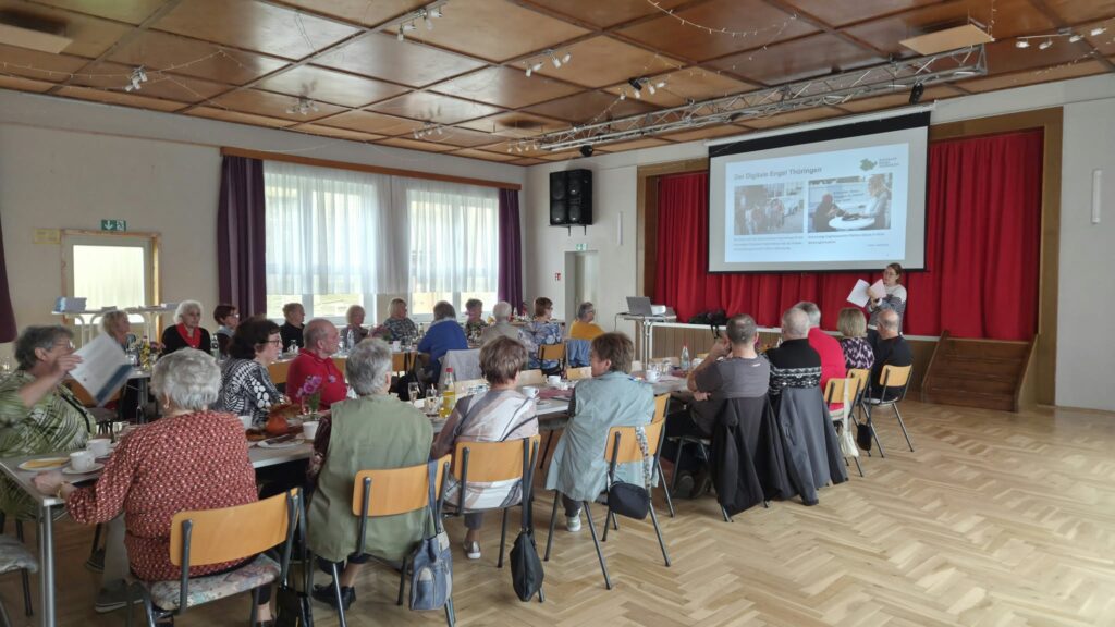 Ein großer Saal mit langen Tischen, an denen ältere Menschen sitzen. Martina steht vor ihnen an einem Redepult. Hinter ihr ist eine Präsentation zu sehen.